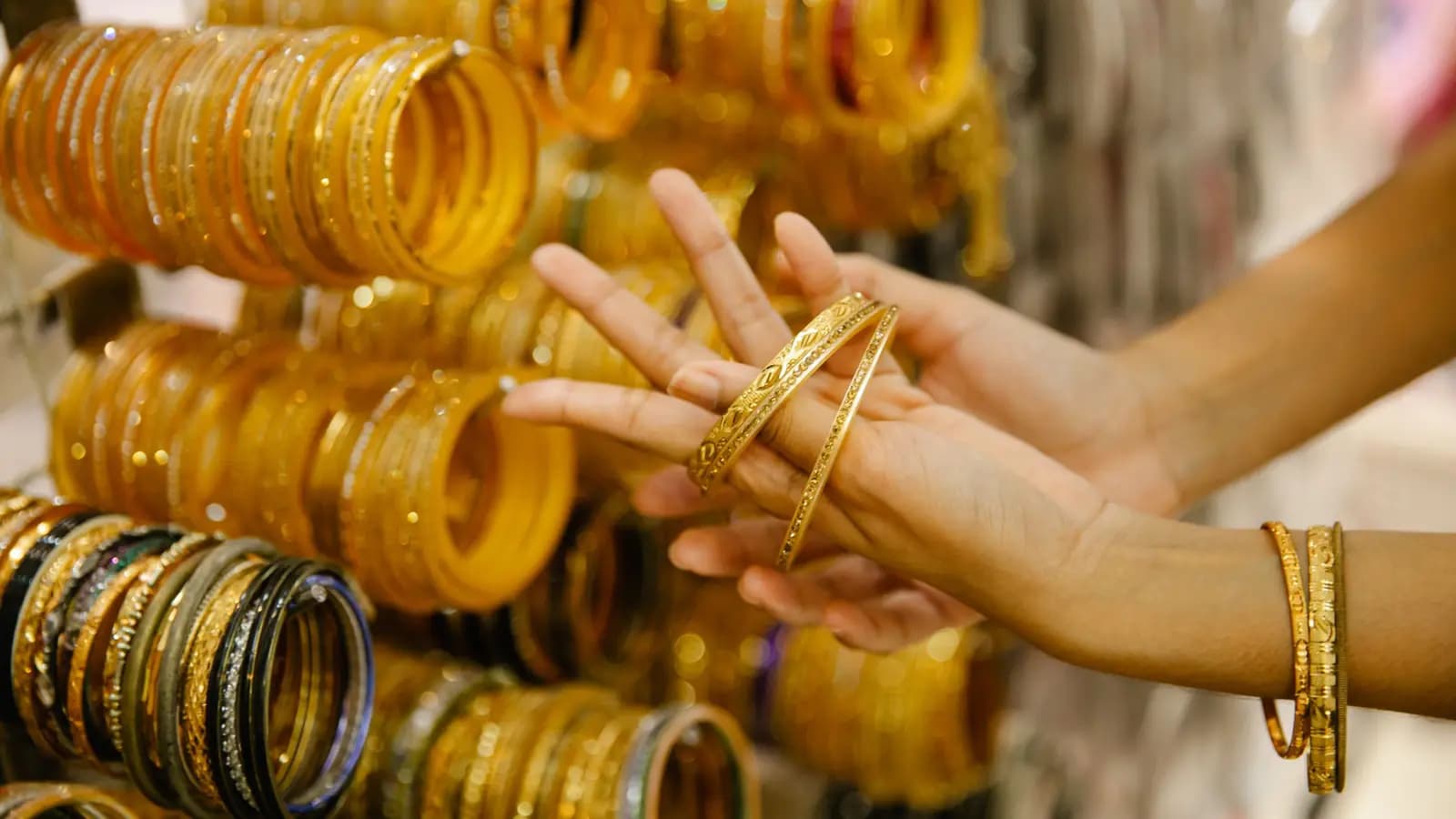 Hands trying on gold bangles in a jewellery shop, with racks of bangles behind